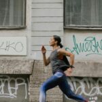 A woman in activewear jogging swiftly past a graffitied wall in an urban setting.