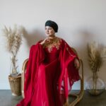 A stylish woman in a red abaya sits indoors, exuding elegance and serenity, surrounded by soft decor.