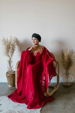 A stylish woman in a red abaya sits indoors, exuding elegance and serenity, surrounded by soft decor.